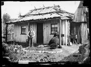 Couple with slab hut, bark-roofed house and garden beds...