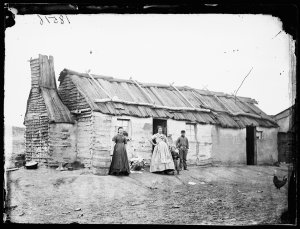 Women and boys outside a wattle and daub house with bar...
