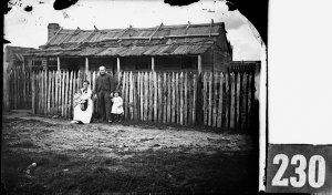 Family in front of weatherboard cottage with bark roof,...