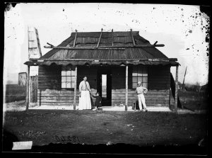 People in front of weatherboard house with bark roof, G...