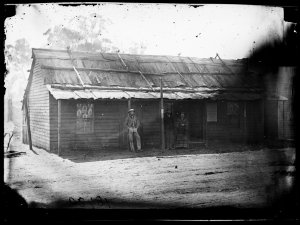 Group in front of weatherboard house with bark roof, Gu...