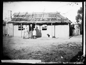 Children in front of whitewashed house with bark roof, ...