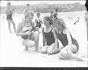 Three young women on Bondi Beach, one playing a ukelele