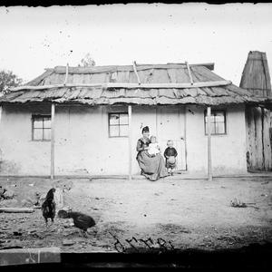 Woman and children with whitewashed house and bark roof...