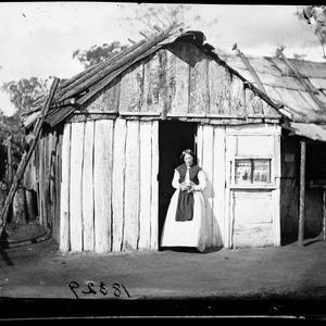 Woman in the door of a partly whitewashed bark house, p...