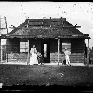 People in front of weatherboard house with bark roof, G...