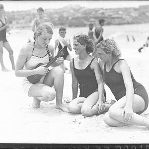 Three young women on Bondi Beach, one playing a ukelele